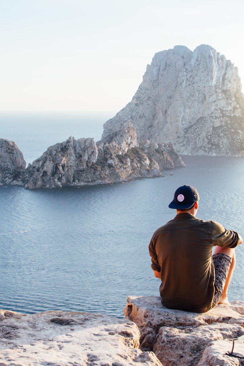 Man enjoys a scenic view of Es Vedrà at sunset from a cliff in San Juan Bautista, providing a perfect summer escape.